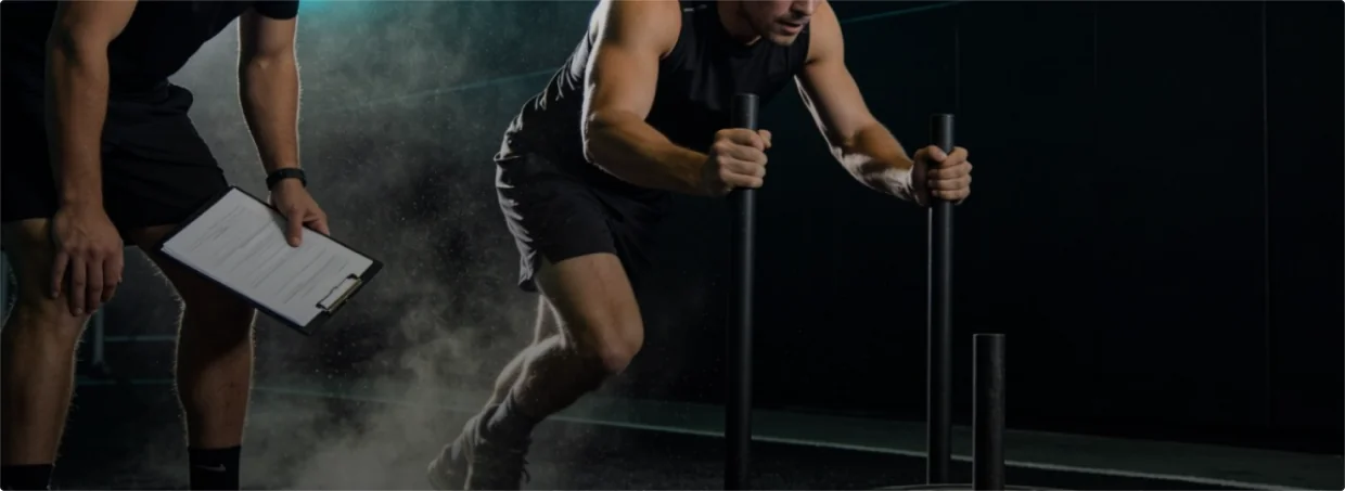Athlete pushing a training sled in a gym while a coach holding a clipboard observes.